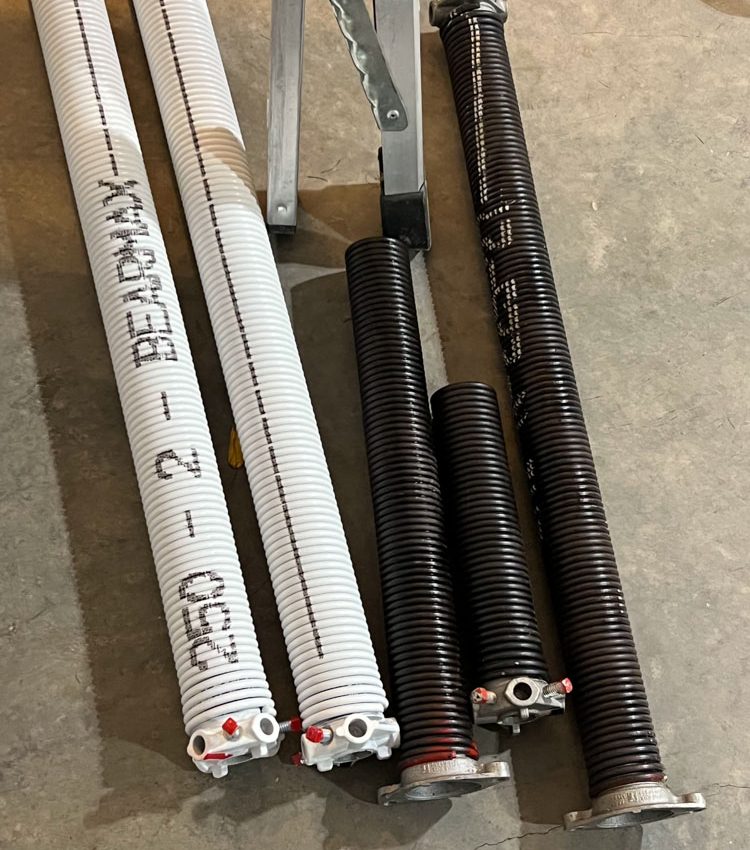 Two new white BEARMAX torsion springs and two old black springs laid out on a concrete floor next to a workbench, prepared for a professional upgrade.