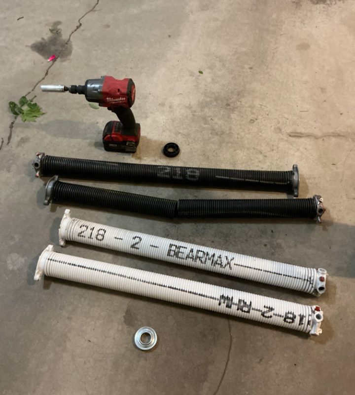Two new white BEARMAX torsion springs and two old black springs laid out on a concrete floor next to a Milwaukee impact driver, ready for replacement.